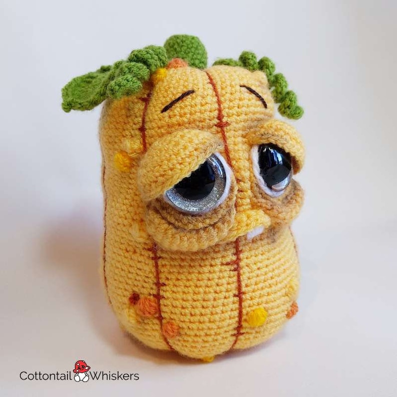 May include: A crocheted pumpkin plush toy with a sad expression, green leaves on top, and brown stitching. The pumpkin is yellow with orange and yellow accents.