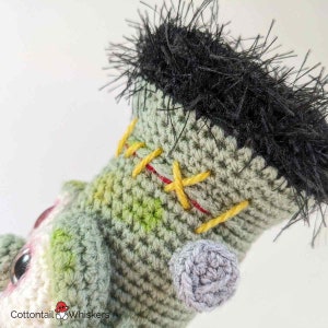 Pode incluir: Close-up de um brinquedo de monstro Frankenstein de croch&ecirc; com fio verde e branco, cabelo de fio preto e costuras amarelas e vermelhas. O monstro tem uma orelha de croch&ecirc; cinza.