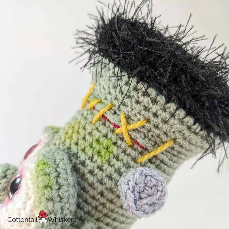 Pode incluir: Close-up de um brinquedo de monstro Frankenstein de croch&ecirc; com fio verde e branco, cabelo de fio preto e costuras amarelas e vermelhas. O monstro tem uma orelha de croch&ecirc; cinza.
