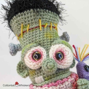 Pode incluir: Close-up de um monstro Frankenstein de croch&ecirc; verde com olhos rosa e branco e costuras amarelas. O monstro est&aacute; usando uma echarpe listrada rosa e branca e segurando um monstro de croch&ecirc; roxo com penas de arco-&iacute;ris.
