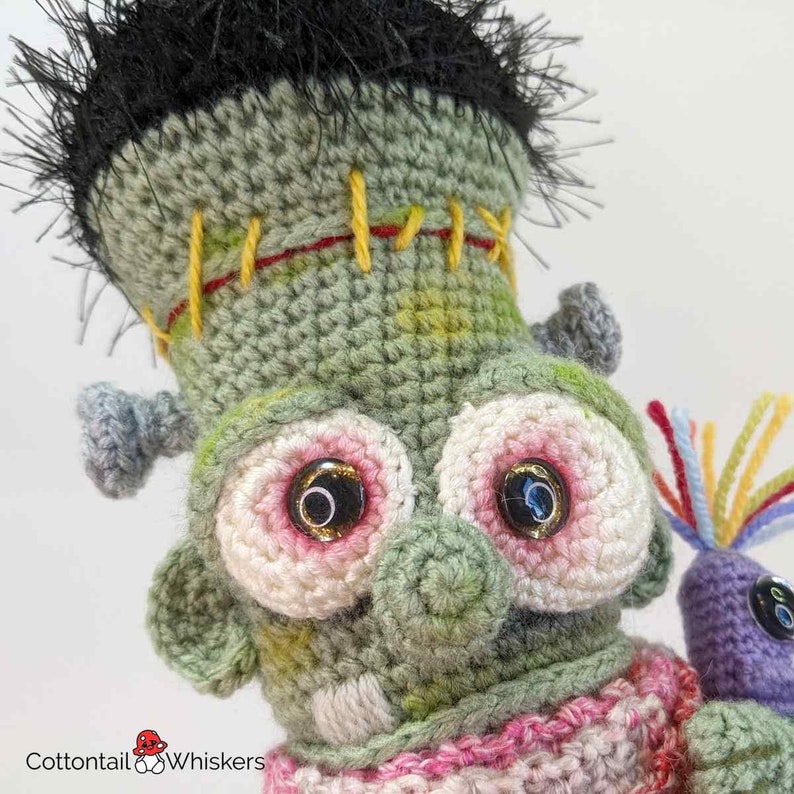 Pode incluir: Close-up de um monstro Frankenstein de croch&ecirc; verde com olhos rosa e branco e costuras amarelas. O monstro est&aacute; usando uma echarpe listrada rosa e branca e segurando um monstro de croch&ecirc; roxo com penas de arco-&iacute;ris.