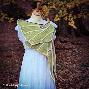 May include: A handmade, green and white crochet shawl with a leaf-like design. The shawl features a scalloped edge, a purple trim, and two circular embellishments. The shawl is displayed on a mannequin, with a white dress in the background.