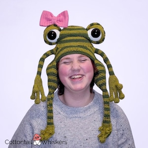 May include: A whimsical, handmade frog hat with large, bulging eyes and long, dangling limbs. The hat is primarily green with darker green stripes, and a pink bow is attached. The hat is designed to be worn on the head.