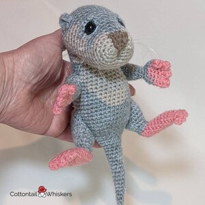 May include: A crocheted otter plush toy with a gray and beige body, pink feet, and a long tail. The otter has a brown nose and black eyes.