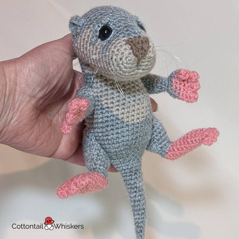 May include: A crocheted otter plush toy with a gray and beige body, pink feet, and a long tail. The otter has a brown nose and black eyes.
