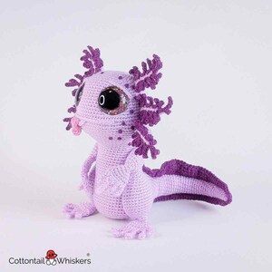 May include: A handmade crochet axolotl in shades of lavender and purple. The plush toy features large, glittery eyes, detailed gills, and a curled tail. The axolotl is posed upright, showcasing its intricate design and soft texture. The logo "Cottontail & Whiskers" is visible.