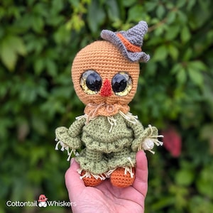 May include: A crocheted scarecrow doll with a pumpkin head, green body, and a gray witch hat. The doll has large, glittery eyes and a red nose.