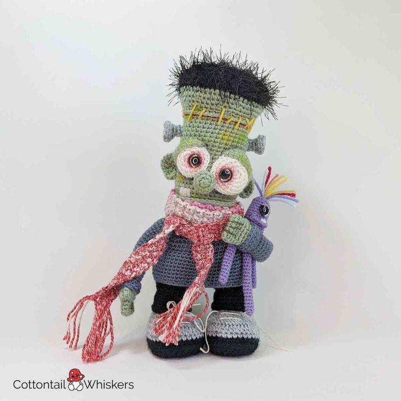 Pode incluir: Um boneco de Frankenstein de croch&ecirc; verde usando uma echarpe vermelha e branca e segurando um boneco de monstro de croch&ecirc; roxo.