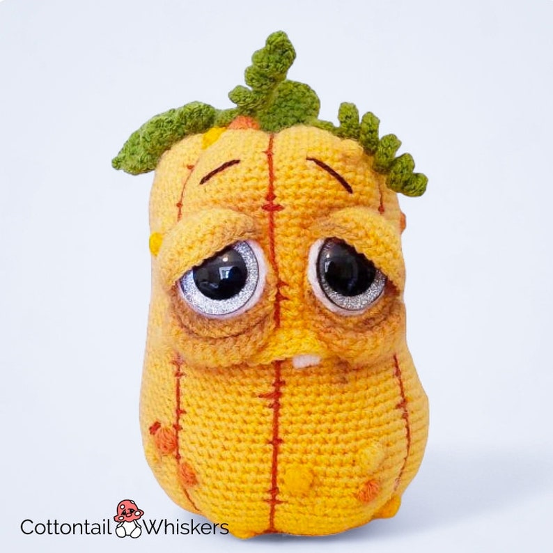 May include: A crocheted pumpkin plush toy with a sad expression. The pumpkin is orange with green leaves on top and has a stitched-on mouth and eyes.