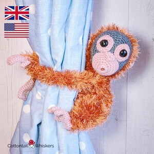 May include: A crocheted monkey curtain tieback in orange and gray with a light blue background. The monkey is holding the curtain tieback with its arms.