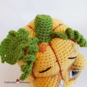 May include: A crocheted pumpkin with a green stem and leaves. The pumpkin has a stitched-on face with closed eyes and a small orange nose.