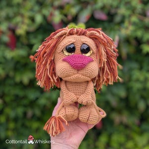 以下が含まれることがあります： 茶色の体、赤いたてがみ、ピンクの鼻をした、かぎ針編みで作られたライオン。ライオンは、金色のキラキラしたアクセントがついた、大きな茶色の目をしています。ライオンは、足を組んで座り、尻尾を後ろに巻いています。