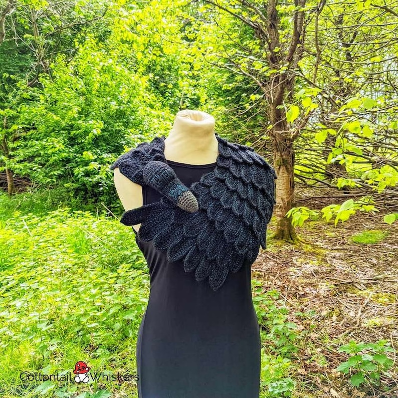 May include: A black crocheted raven cowl with a detailed wing design. The cowl is worn on a mannequin against a green background.