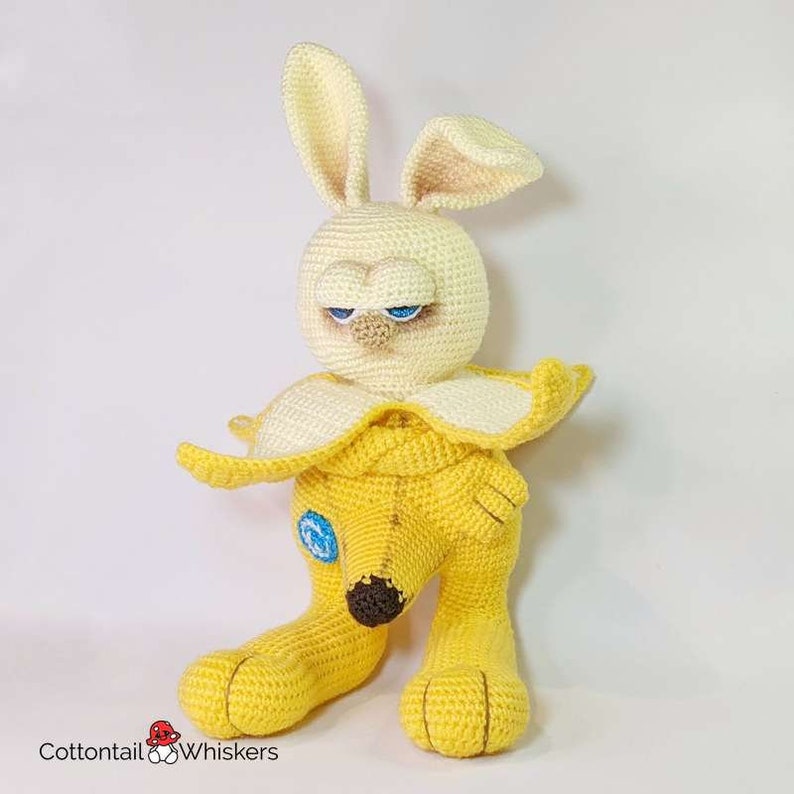 May include: A crocheted stuffed toy rabbit wearing a banana costume. The rabbit has yellow fur, a brown nose, and blue eyes. The banana costume is yellow and has a brown stem.