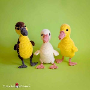 May include: Three crocheted ducklings standing side by side. The left duckling is yellow and brown with a dark gray beak and feet. The center duckling is cream-colored with a pink beak and feet. The right duckling is yellow with a pink beak and feet.