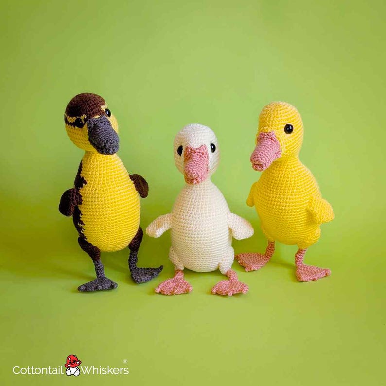 May include: Three crocheted ducklings standing side by side. The left duckling is yellow and brown with a dark gray beak and feet. The center duckling is cream-colored with a pink beak and feet. The right duckling is yellow with a pink beak and feet.