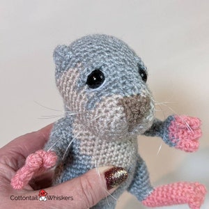 May include: A crocheted gray and pink stuffed animal with black eyes and a brown nose. The animal has long whiskers and is being held by a hand.