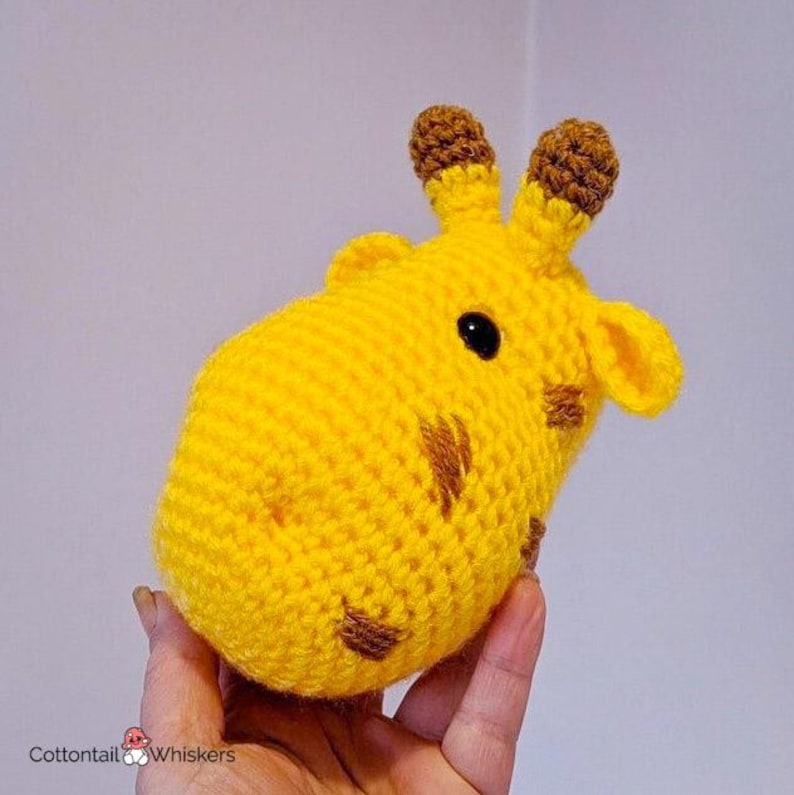 Puede incluir: Un juguete de peluche de jirafa tejido a crochet con orejas y manchas marrones. La jirafa es amarilla y tiene ojos negros.