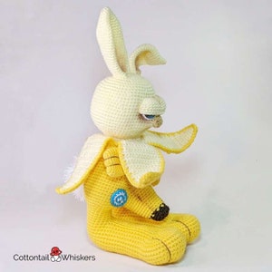 May include: A crocheted banana-themed bunny plush toy with a yellow body, white fur, and a blue eye. The bunny is sitting with its legs crossed and has a grumpy expression.