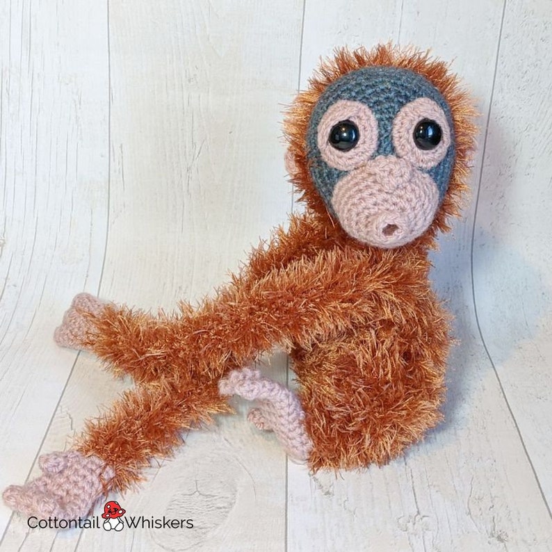 May include: A crocheted orangutan plush toy with a light brown, fuzzy body, a gray face, and large black eyes. The toy is sitting on a white wooden surface.