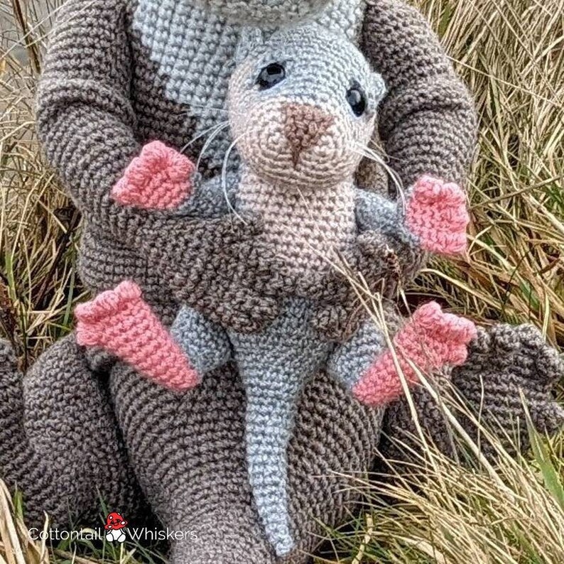 May include: A crocheted gray and brown otter holding a smaller crocheted gray otter with pink feet. The larger otter is wearing a gray and brown crocheted sweater. The smaller otter is sitting in the larger otter's arms. The background is a blurry image of brown grass.