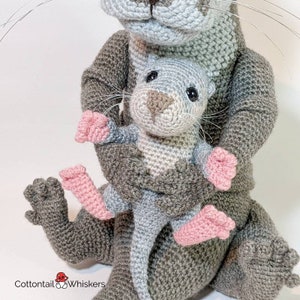 May include: A gray crocheted otter holding a smaller gray crocheted otter with pink feet. The larger otter has a light gray face and a long tail.