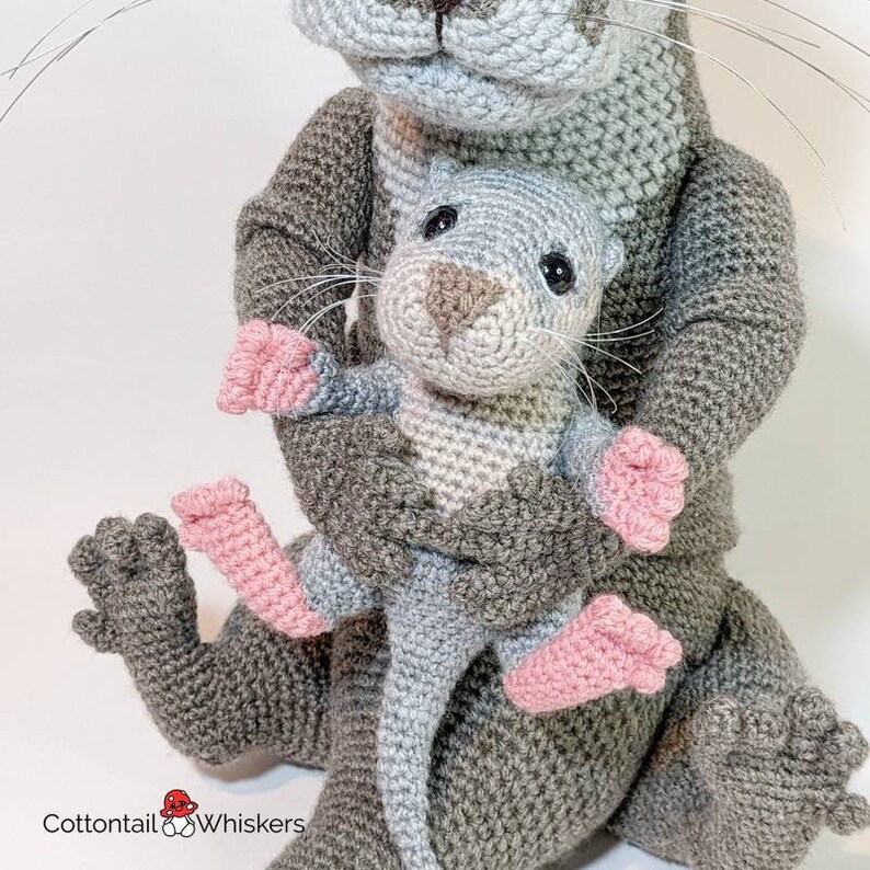 May include: A gray crocheted otter holding a smaller gray crocheted otter with pink feet. The larger otter has a light gray face and a long tail.