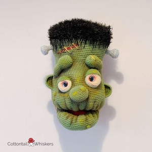May include: Green crocheted Frankenstein monster puppet with black hair, stitched scars, and a stitched mouth. The puppet has large, round eyes with white pupils.
