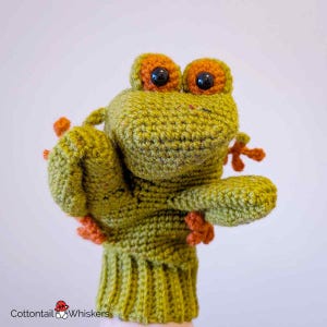May include: A handmade green and orange crochet frog puppet. The puppet has large orange eyes with black pupils, orange feet, and a green body. The puppet is made of yarn and is designed to fit over a hand.
