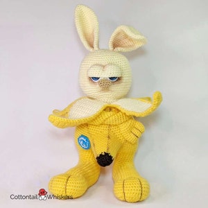 May include: A crocheted stuffed toy shaped like a banana with a bunny face. The toy is yellow and has blue eyes. The toy is sitting on a white surface.
