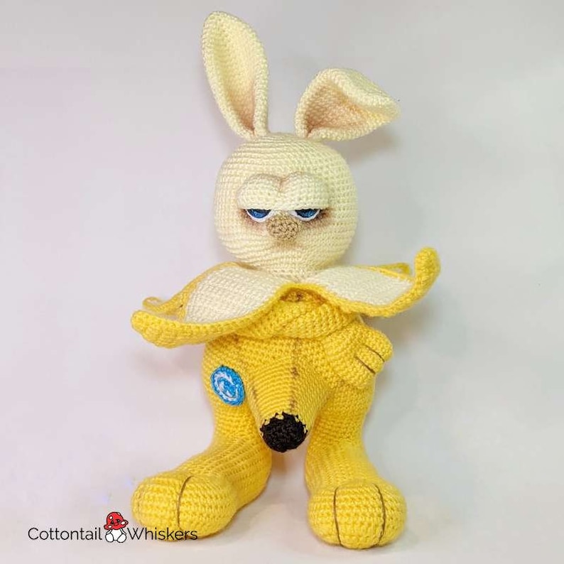 May include: A crocheted stuffed toy shaped like a banana with a bunny face. The toy is yellow and has blue eyes. The toy is sitting on a white surface.
