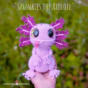 May include: A crocheted axolotl toy in shades of pink and purple, with large, glittery eyes and a protruding pink tongue. The axolotl has detailed gills and limbs. The text "SPRINKLES THE AXOLOTL" is above the toy.