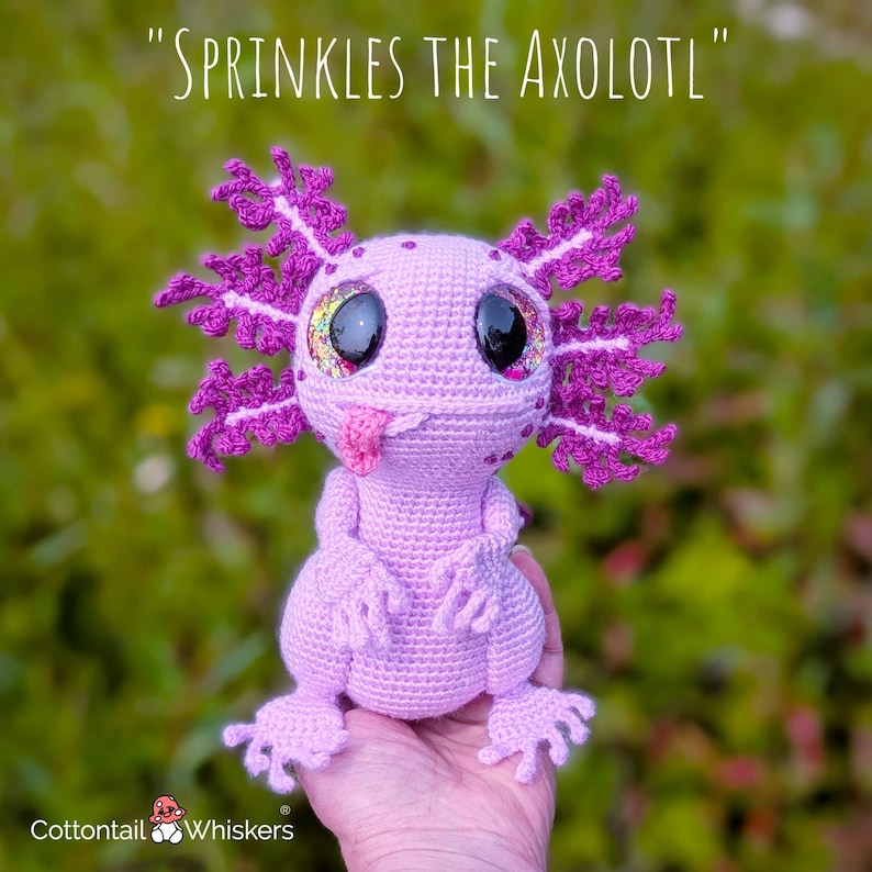 May include: A crocheted axolotl toy in shades of pink and purple, with large, glittery eyes and a protruding pink tongue. The axolotl has detailed gills and limbs. The text "SPRINKLES THE AXOLOTL" is above the toy.