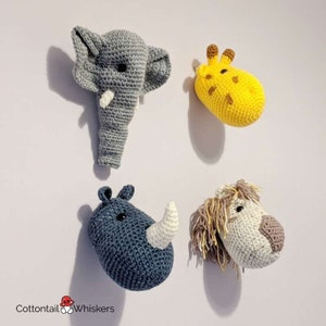 Puede incluir: Cuatro cabezas de animales de crochet, incluyendo un elefante, una jirafa, un rinoceronte y un le&oacute;n, se muestran en una pared blanca. El elefante es gris, la jirafa es amarilla, el rinoceronte es azul y el le&oacute;n es blanco con una melena marr&oacute;n.