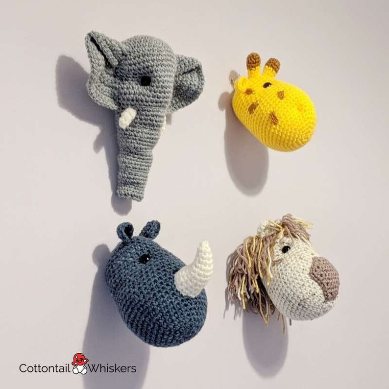 Puede incluir: Cuatro cabezas de animales de crochet, incluyendo un elefante, una jirafa, un rinoceronte y un le&oacute;n, se muestran en una pared blanca. El elefante es gris, la jirafa es amarilla, el rinoceronte es azul y el le&oacute;n es blanco con una melena marr&oacute;n.