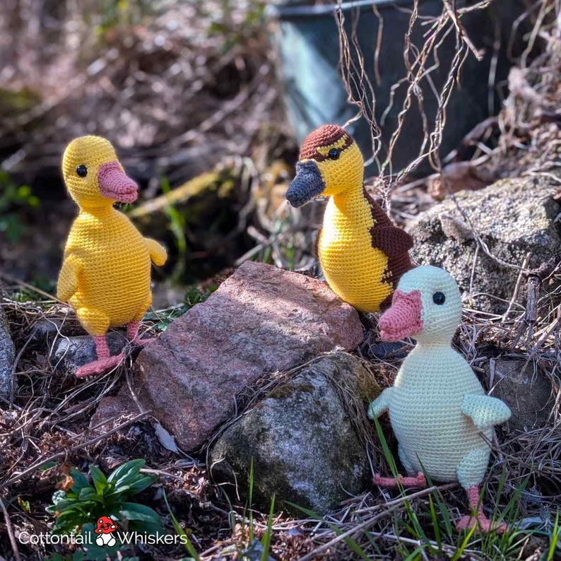 May include: Three crocheted ducklings in yellow, tan, and cream colors are arranged on a rocky surface. The ducklings have pink beaks and feet, with one duckling featuring a brown head. The image includes the text "Cottontail Whiskers".