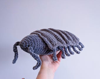 May include: Grey crocheted plush toy shaped like a pill bug. The toy has segmented body parts and small legs.