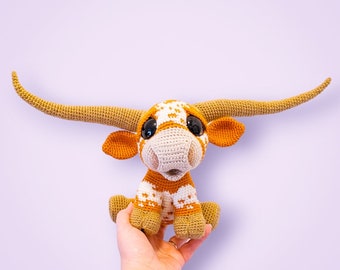Puede incluir: Un juguete de peluche de crochet de un buey Texas Longhorn con grandes cuernos marrones, manchas blancas y naranjas y grandes ojos marrones. El juguete está sentado en una mano.