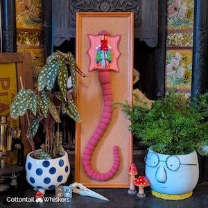 May include: A pink crocheted anatomical art piece in a frame, mounted on a corkboard. The artwork is displayed with potted plants, including a polka-dotted planter and a face-shaped pot. Two small mushroom figurines and a bird skull are also present.
