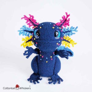 May include: A handmade, dark blue crochet axolotl with colorful accents. The axolotl features pink, yellow, and blue frills, with small colorful dots on its head and body. It has large, expressive eyes and small arms and legs.