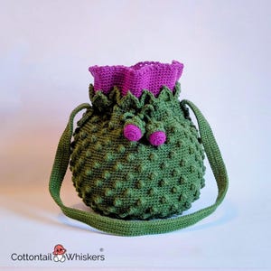May include: A handmade green crochet bag with a textured, bumpy surface. The bag features a purple ruffled top and two small purple accents. It has a long green strap for carrying.