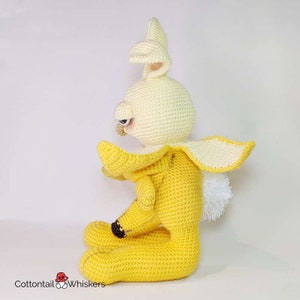 May include: A crocheted banana-shaped bunny plush toy with a white face and yellow body. The bunny is sitting with its head tilted to the side and its arms outstretched.