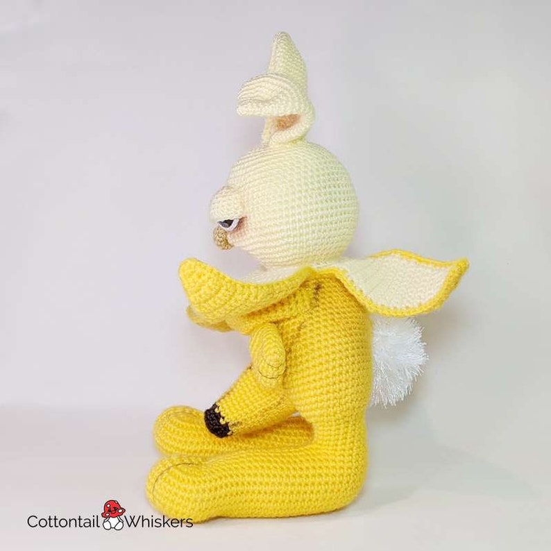 May include: A crocheted banana-shaped bunny plush toy with a white face and yellow body. The bunny is sitting with its head tilted to the side and its arms outstretched.