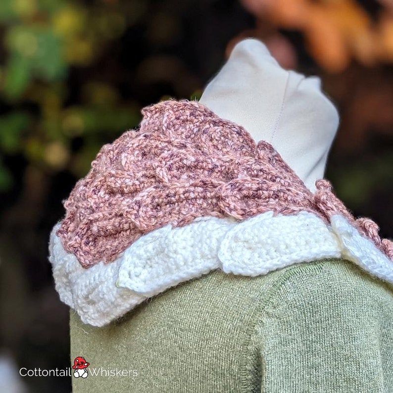 Pu&ograve; includere: Un collo a scialle in crochet rosa e bianco con un design testurizzato. Il collo &egrave; indossato su un manichino con un maglione verde.