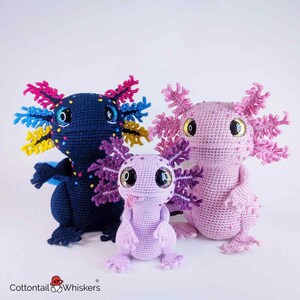 May include: Three crocheted axolotl plushies in varying sizes. The largest is navy blue with colorful gills, the medium is pink, and the smallest is lavender. All have large, round eyes and detailed features.