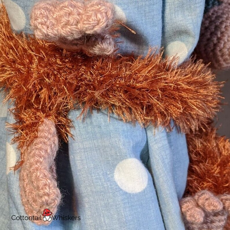 May include: A close-up of a crocheted monkey wearing a blue and white polka dot dress. The monkey has a fluffy orange tail and pink crocheted arms and legs.