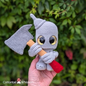 Puede incluir: Un juguete de crochet gris con un sombrero puntiagudo y ojos grandes que sostiene un hacha roja y naranja. El juguete está siendo sostenido en la mano de una persona.