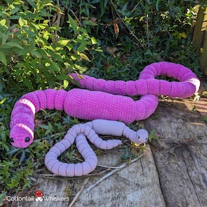 Puede incluir: Dos peluches de gusano de crochet, uno grande de color rosa y otro más pequeño de color gris, están tumbados sobre una superficie de madera cubierta de follaje verde.