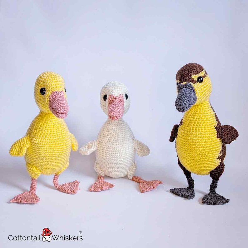 May include: Three crocheted ducklings in yellow, white, and brown. The ducklings have pink beaks and feet, and black eyes. The ducklings are handmade and appear to be toys or decorative items.