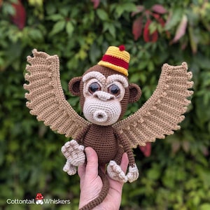 May include: A brown crocheted monkey with large eyes and a red and yellow hat. The monkey has wings and is holding a small whistle in its mouth.